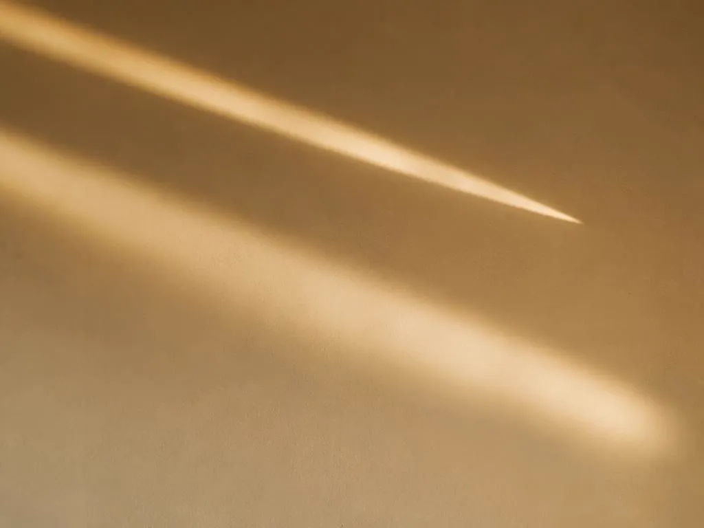 Two beams of warm light crossing a cream wall, one ending in a clean point and one fading softly